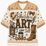 A short-sleeve V-neck t-shirt with a cream and tan base, featuring a detailed graphic print on the front. The graphic includes the text 'She's a ART I can Tell' in leopard-print outlined letters, surrounded by artistic and western-themed illustrations such as a paint palette, brushes, cowboy boots, bows, stars, and daisies.