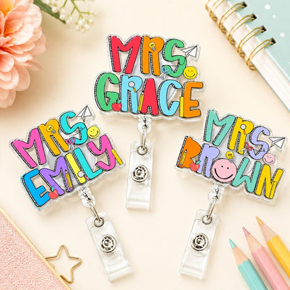 Colorful name badge holders with 'Mrs. Grace', 'Mrs. Emily', and 'Mrs. Brown' on a desk with stationery items.