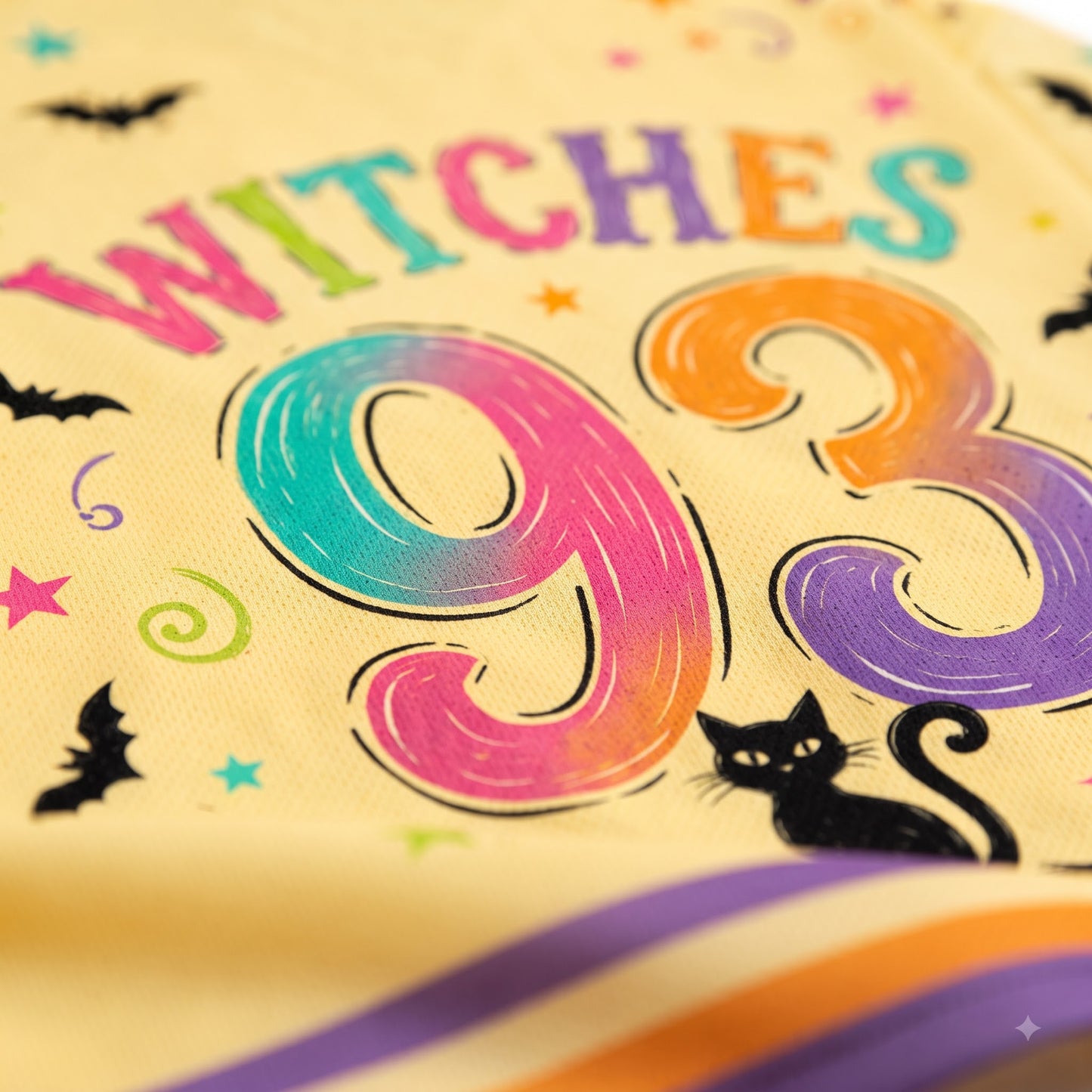 A close-up of a fabric product featuring a colorful 'WITCHES 93' Halloween design on a cream-colored background, with black bats, stars, swirls, and a black cat illustration.