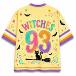 A pale yellow short-sleeve T-shirt shown from the back, decorated with colorful Halloween graphics including the words “WITCHES 93” in rainbow lettering, black bats, stars, swirls, a black cat on a broom, and purple, orange, and white striped sleeve accents.