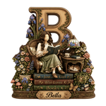 A decorative figurine of a woman sitting in a chair and reading a book, styled with a large ornate letter B, flowers, books, a small table with a lamp and teacup, mushrooms, and a plaque reading 'Bella'.