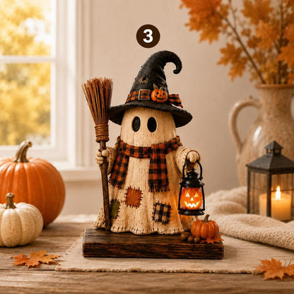 A Halloween-themed decorative ghost figure made from a wood slice, wearing a black witch hat with a small orange pumpkin accent, a red-and-black plaid scarf, and holding a broom and a glowing lantern. The figure stands on a dark wooden base on a tabletop, surrounded by pumpkins, autumn leaves, a candle, and a vase with dried leaves in a softly lit room.
