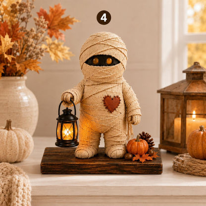 A mummy-shaped decorative figurine wrapped in beige faux-fur or fabric, with black eyes, holding a glowing black lantern. It stands on a dark wooden base with small pumpkins, a pinecone, and autumn leaves, set on a tabletop with fall decor in the background.