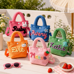 Five personalized corduroy handbag charms in pink, blue, green, orange, and light pink, each with embroidered names and cute character accents, displayed on white wooden steps outdoors with strawberries and pink sunglasses nearby.
