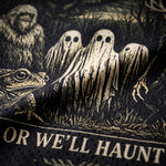 Close-up of a black mesh fabric printed with a spooky Halloween graphic featuring three ghost figures, a frog, a Yeti, and partial text reading 'OR WE'LL HAUNT'.