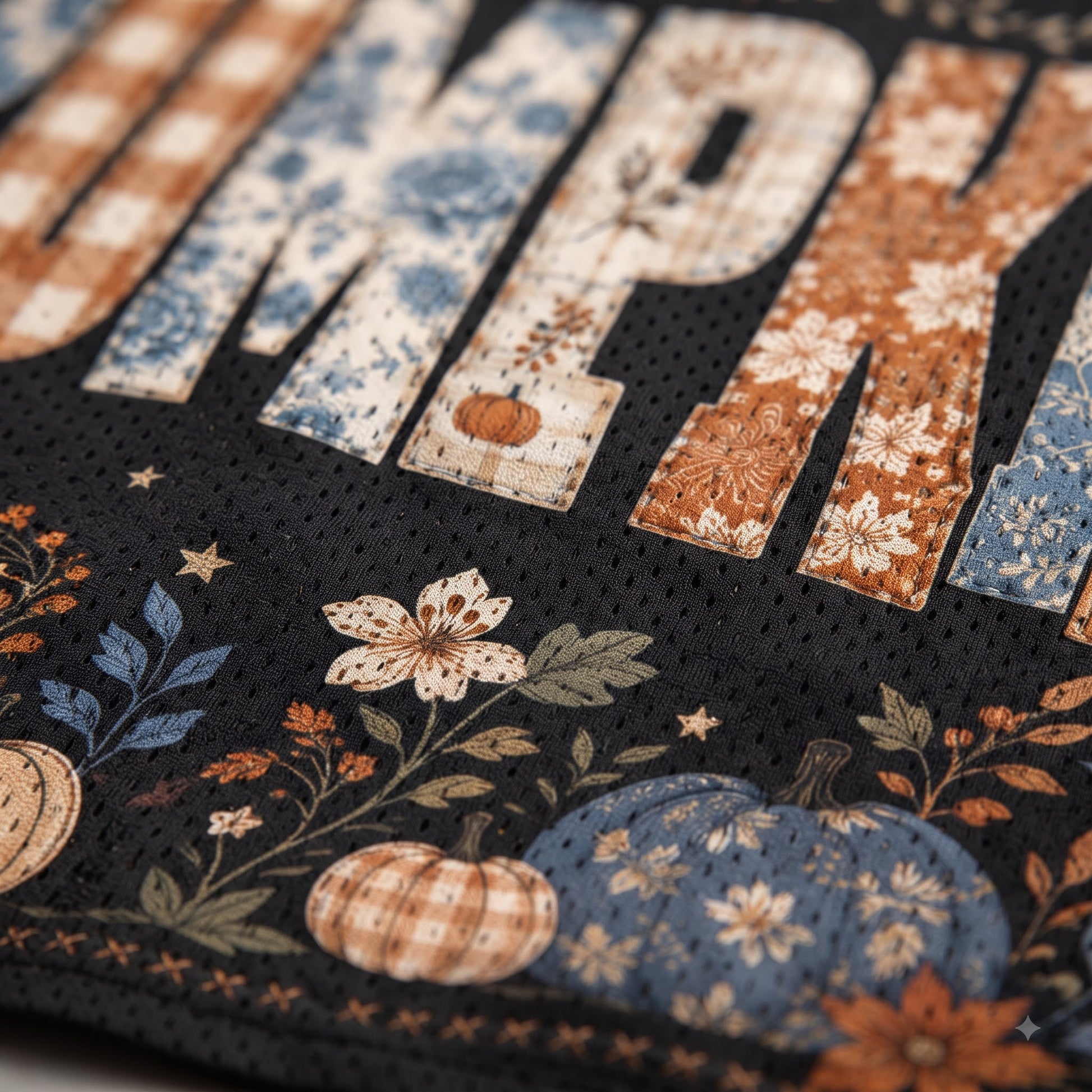 Close-up of a black mesh fabric with a fall harvest pattern featuring large floral and plaid letters, pumpkins, stars, and leaves in blue, orange, beige, and white.