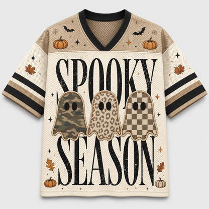 A short-sleeve V-neck jersey-style shirt with a beige and cream Halloween graphic. The front reads 'SPOOKY SEASON' in large black letters over three ghost silhouettes in camouflage, leopard print, and checkerboard patterns, with pumpkins, bats, stars, and leaves on a light gray background.