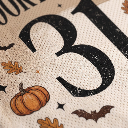 Close-up of a textured cream fabric print featuring a large distressed black number 3, an orange pumpkin, brown leaves, bats, and small stars in a Halloween theme.